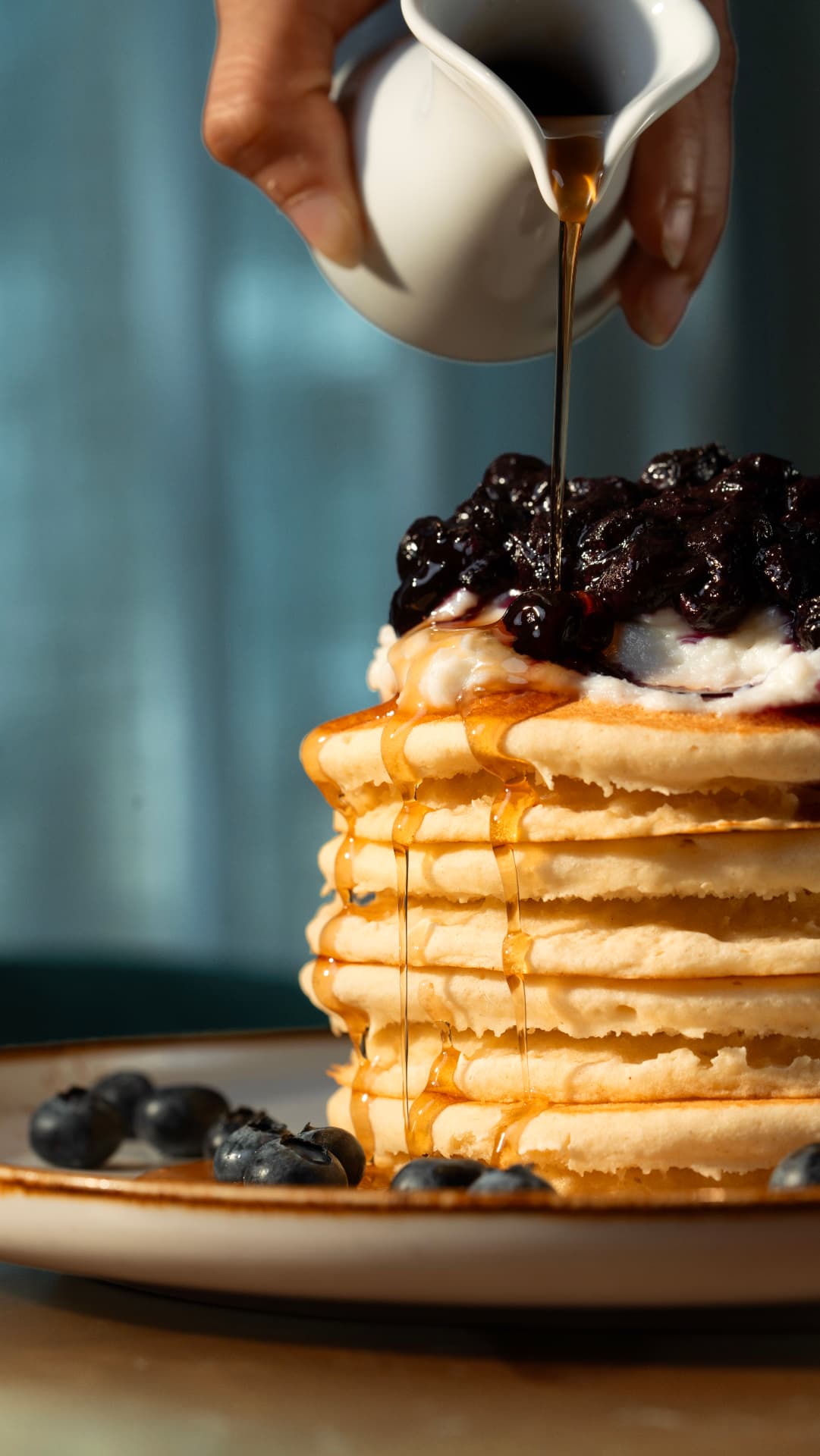 Maple syrup poured from a jug over a tall stack of buttermilk pancakes topped with blueberry compote at SOLENA