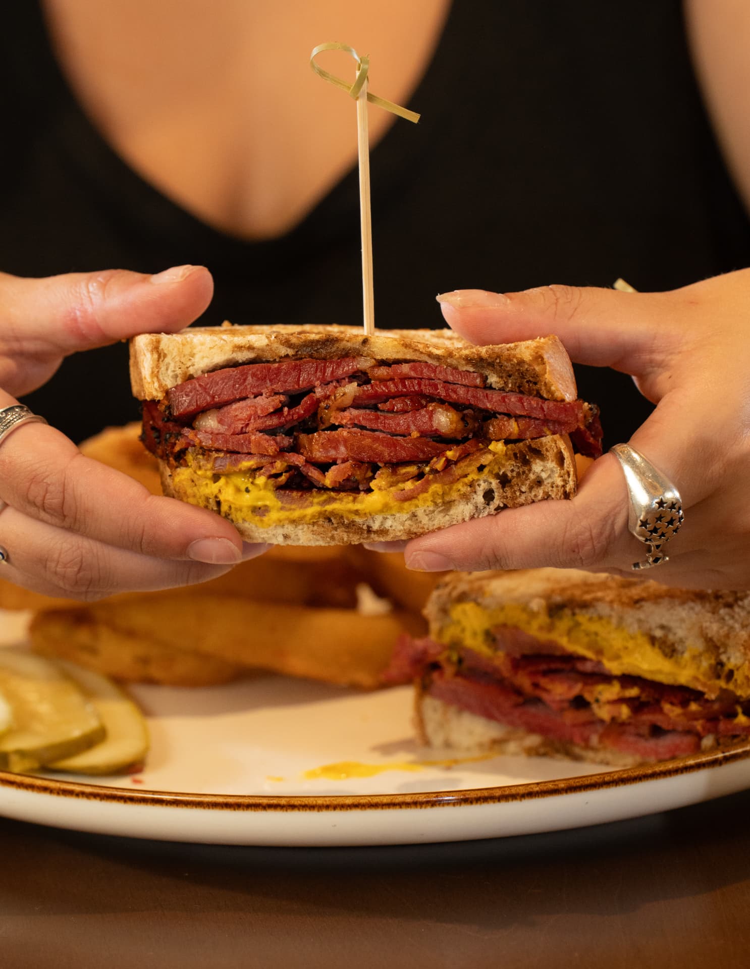 Guest holding a toasted pastrami sandwich with mustard, served with fries and pickles at SOLENA
