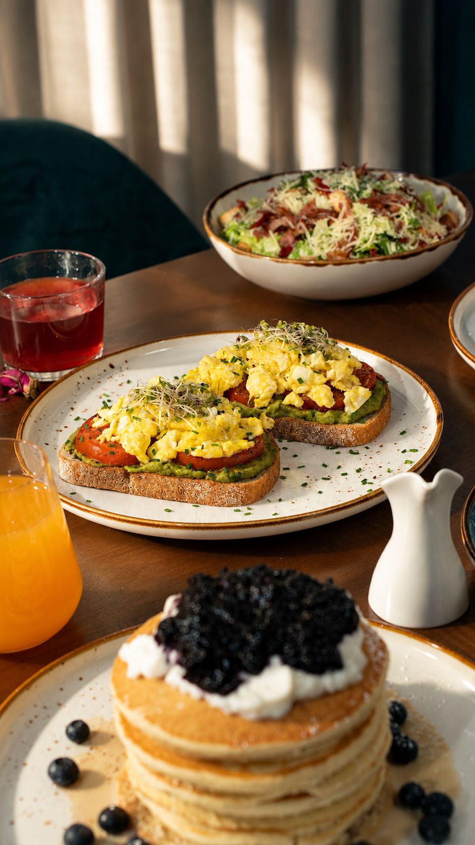 Avocado toast with scrambled eggs and sprouts beside a Caesar salad, mimosa and blueberry pancakes at SOLENA