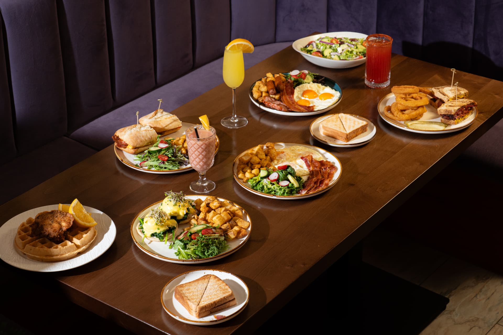 Full brunch spread across a walnut table in SOLENA's violet velvet booth — benedicts, pastrami sandwich, chicken and waffles, mimosa and caesars