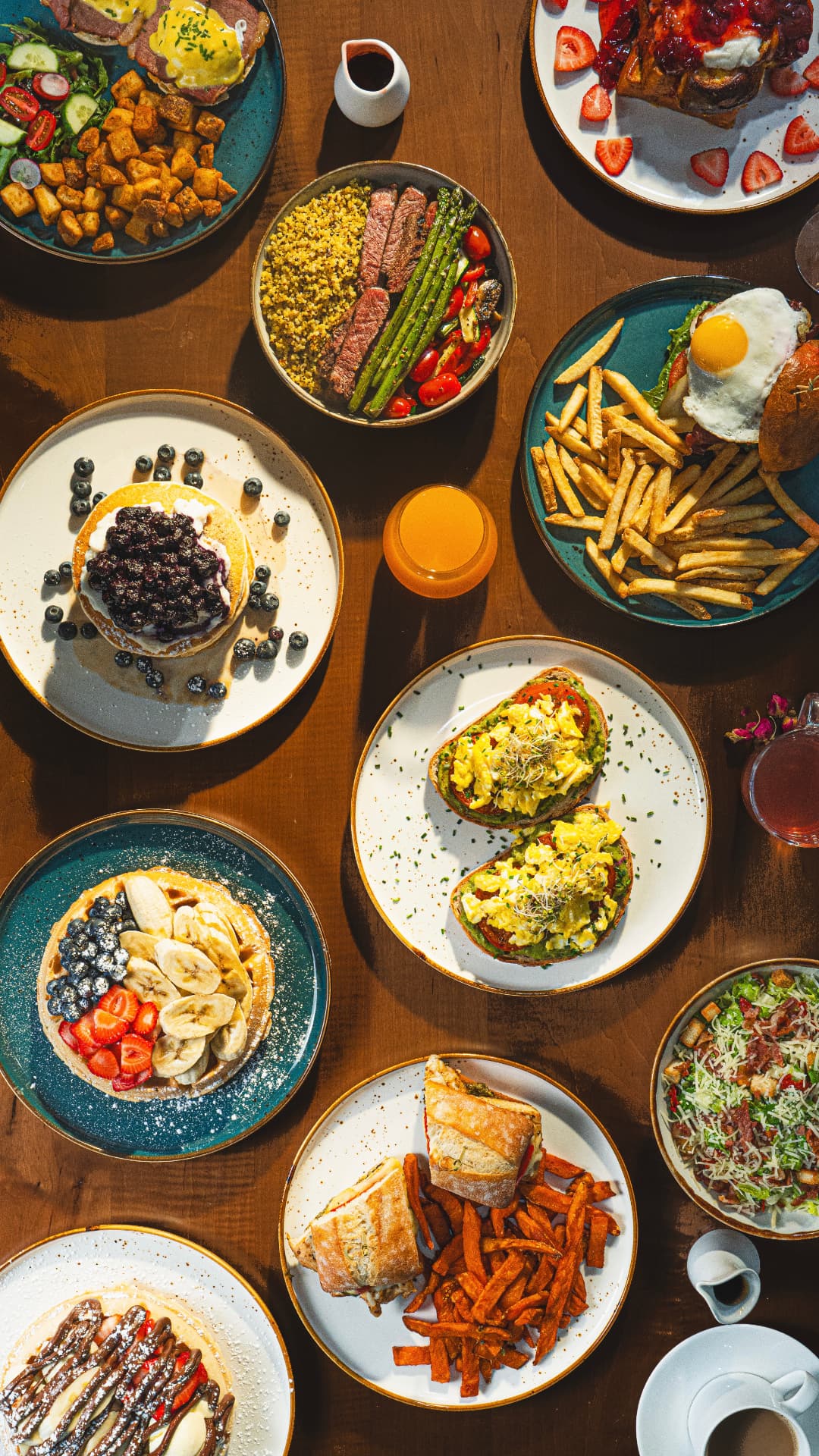 Overhead table spread at SOLENA — benedicts, steak bowl, blueberry pancakes, avocado toast, waffles and burgers with fries
